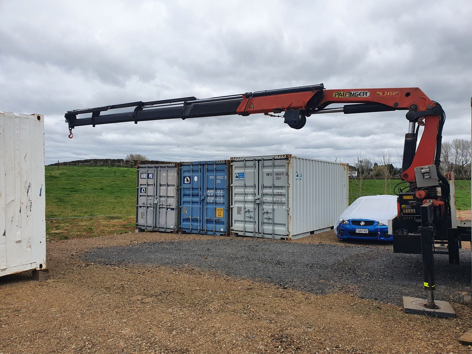 The Palfinger crane boom extended across a gravel yard, outrigger down, shipping containers behind