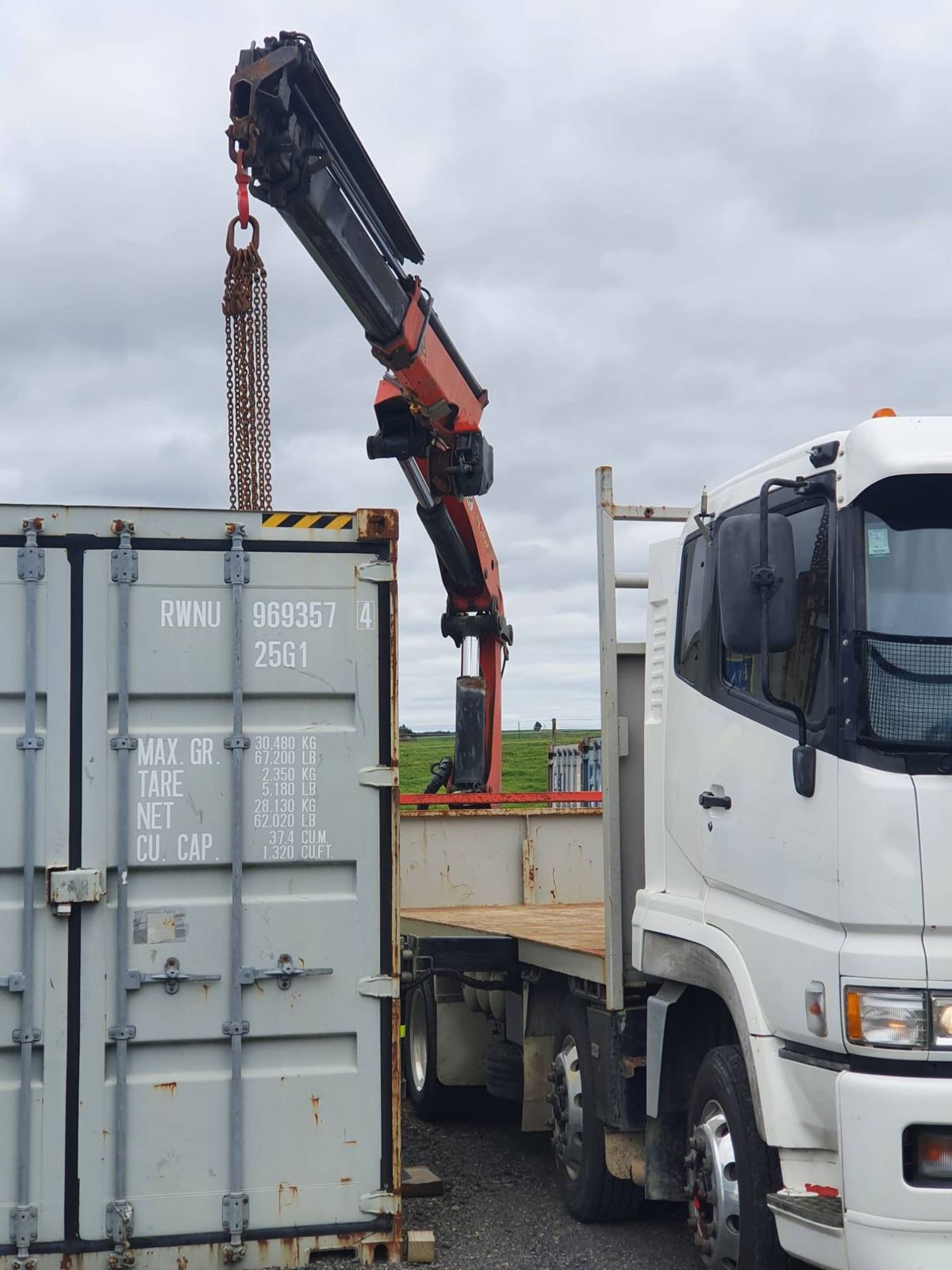 Crane boom raised with lifting chains hanging over the end of a shipping container, truck cab alongside