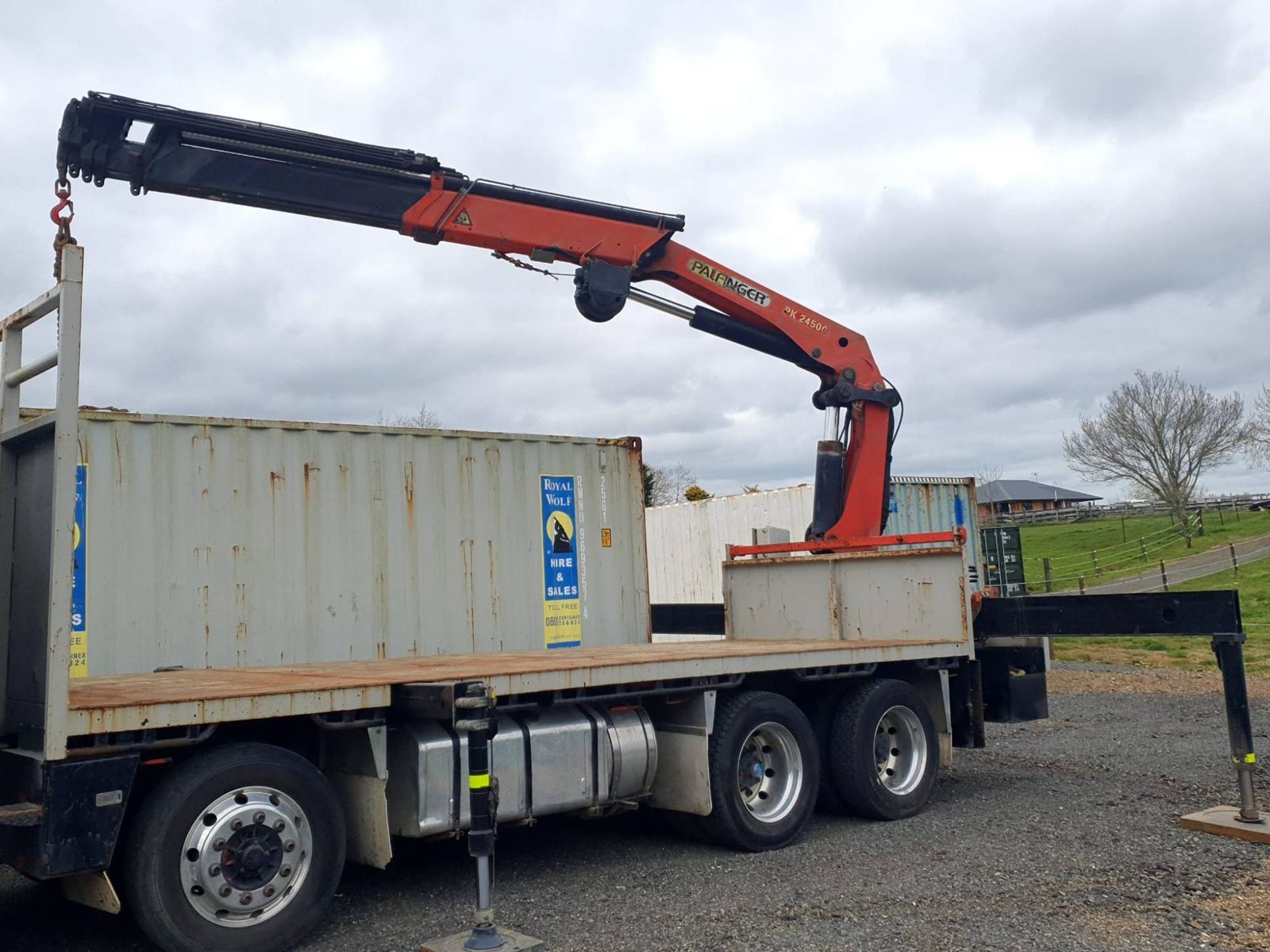 White eight-wheeler crane truck with a Palfinger crane at the rear and a 20 ft shipping container on its flat deck, outrigger down on gravel
