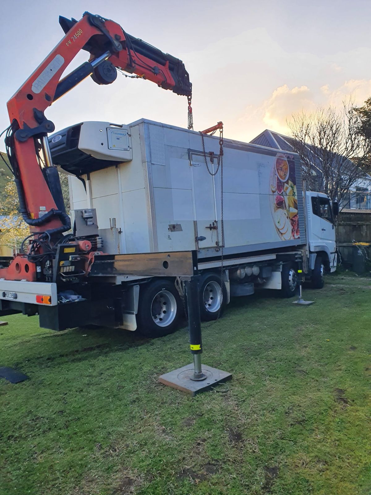 The Palfinger crane lifting a refrigerated truck body with chains, outriggers down on pads on a lawn